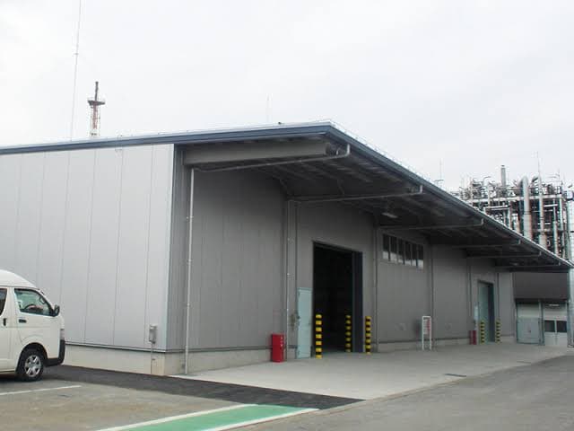 グレーの波板外装の倉庫・処理施設。積込スペースと大型シャッターを備えた拠点の外観。
